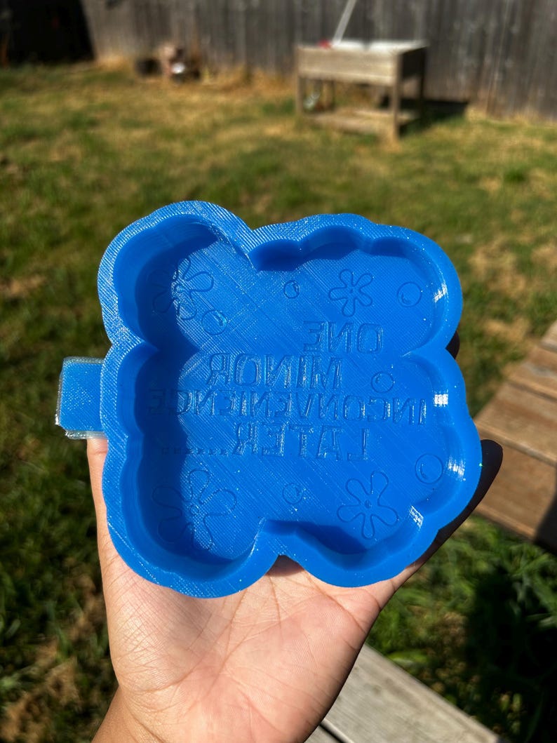 Op de afbeelding: Een helderblauwe, bloemvormige mal met de tekst "ONE MINUTE LATER!" en decoratieve bloemelementen. De mal wordt in een hand gehouden tegen een achtergrond van groen gras en een houten hek.