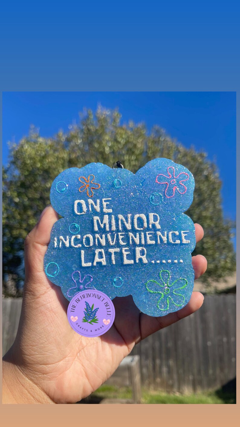 Op de afbeelding: Een lichtblauwe, glinsterende luchtverfrisser met de tekst "ONE MINOR INCONVENIENCE LATER....." en bloemmotieven. De luchtverfrisser wordt in een hand gehouden tegen een blauwe hemel.