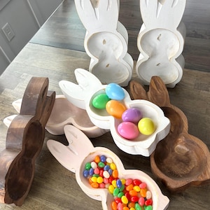 May include: A set of wooden bunny-shaped bowls, two white, one pink, and two brown. The white bowls are filled with colorful plastic Easter eggs, the pink bowl is filled with colorful jelly beans, and the brown bowls are empty.