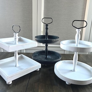 May include: Four decorative tiered trays in the image. Two are white with square and oval tiers, and one is black with two round tiers. Each tray has a handle and is made of wood.
