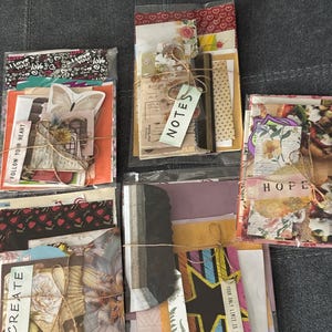 May include: Several clear plastic bags filled with assorted paper ephemera, tied with twine. The bags contain patterned papers, tags with text like "NOTES", "CREATE", and "HOPE", and decorative elements. The overall aesthetic is vintage and artistic.