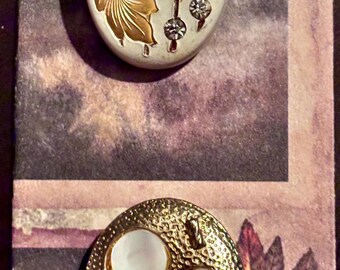 May include: Two oval-shaped buttons. The top button is off-white with gold floral designs and clear crystal accents. The bottom button is gold-toned with a textured surface and four mother-of-pearl inlays. Both buttons are set against a patterned background.