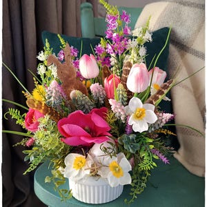May include: A white ceramic vase filled with a spring floral arrangement. The arrangement includes pink tulips, white daffodils, pink and purple flowers, and green foliage. A wooden bunny figurine is nestled in the centre of the arrangement.