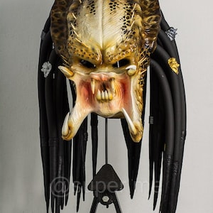 Puede incluir: Una detallada máscara de Depredador con una cabeza texturizada de color amarillo dorado y marrón, dientes afilados y rastas negras. La máscara se exhibe sobre un soporte de metal negro, mostrando el intrincado diseño y las amenazantes características del cazador alienígena.