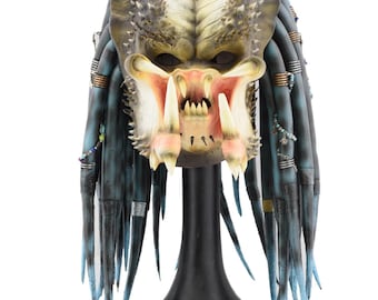 May include: A detailed Predator mask replica, featuring a textured, weathered appearance with shades of green, tan, and blue. The mask has prominent mandibles, tusks, and dreadlocks, mounted on a black display stand. A collectible item.