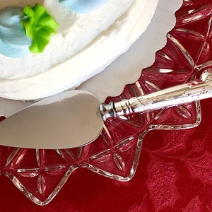 以下が含まれることがあります： 装飾的なハンドルが付いた銀製のケーキサーバーが、透明なガラス製のケーキプレートの上に置かれています。プレートは赤いテーブルクロスの上にあります。