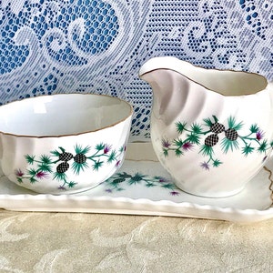 Puede incluir: Un juego de porcelana blanca que incluye una jarra para crema, un azucarero y una bandeja. El juego presenta un diseño de cardo en verde, morado y negro. Los bordes de la bandeja y el cuenco son festoneados y dorados.
