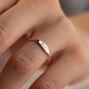 May include: A silver ring featuring a small, clear gemstone set within a curved, oval shape. The ring is worn on a finger, demonstrating its delicate and minimalist design. The polished surface of the ring reflects light, emphasising its elegant simplicity.