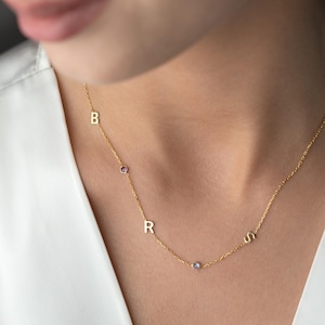 May include: A delicate gold necklace with the letters B, R, and S, interspersed with small purple gemstones. The necklace is worn against a white background, highlighting the gold chain and the gemstones' color.