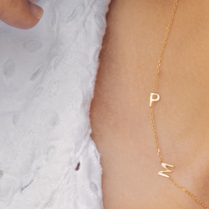 May include: A gold chain necklace with a small letter "P" and a small letter "M" charm. The necklace is worn by a person with a white shirt.