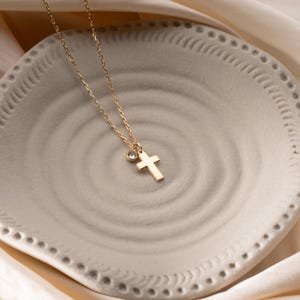 Op de afbeelding: Een goudkleurige ketting met een kleine kruisvormige hanger en een bedel met een heldere edelsteen. De ketting wordt gepresenteerd op een lichtgrijze, getextureerde plaat met een decoratieve rand, tegen een crèmekleurige stof.
