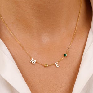 May include: A gold-toned chain necklace featuring the letters M and E in a polished finish, accented by a small round yellow gemstone between the letters and a round green gemstone positioned to the right of the letter E on the delicate chain.