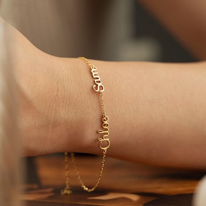 May include: A gold-colored bracelet with the names "Sam" and "Chloe" in cursive script. The delicate chain bracelet is worn on a wrist, showcasing the personalized jewelry.