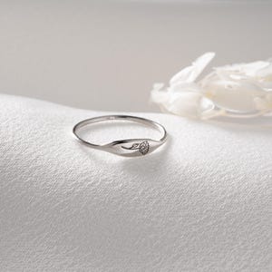 May include: A thin silver ring featuring a small, flat, oval-shaped face engraved with a delicate, single-line drawing of a flower. The band is slender and polished, showcasing a metallic finish that reflects light across its smooth, curved surface.