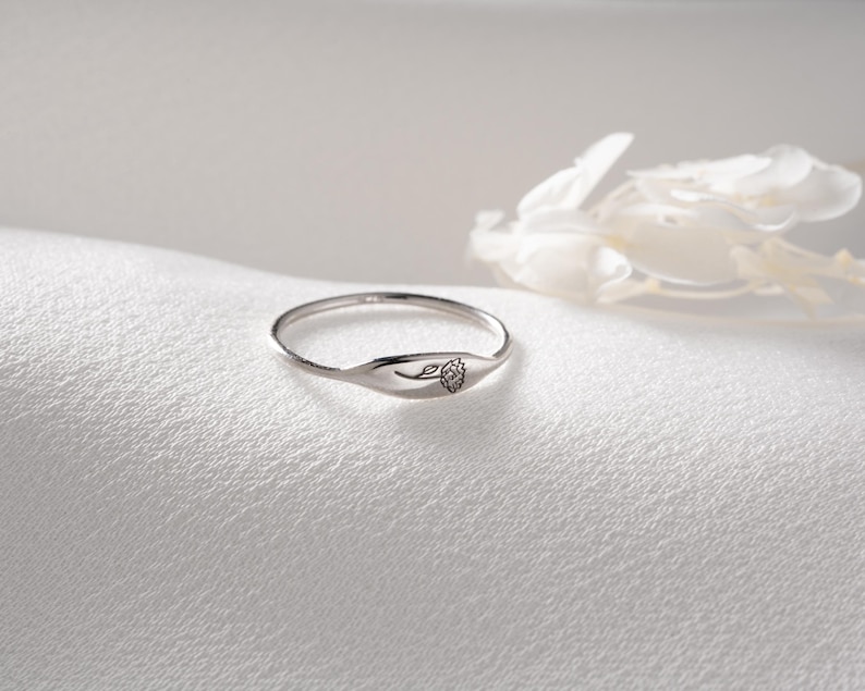 May include: A thin silver ring featuring a small, flat, oval-shaped face engraved with a delicate, single-line drawing of a flower. The band is slender and polished, showcasing a metallic finish that reflects light across its smooth, curved surface.