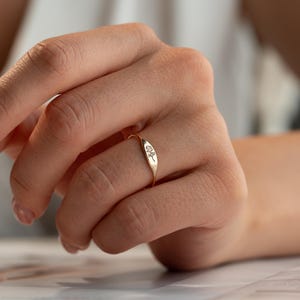 May include: A gold-toned signet ring featuring a small, engraved floral design on the flat, oval-shaped face. The band is thin and circular, with the metal surface displaying a polished finish that reflects light across the surface of the ring.