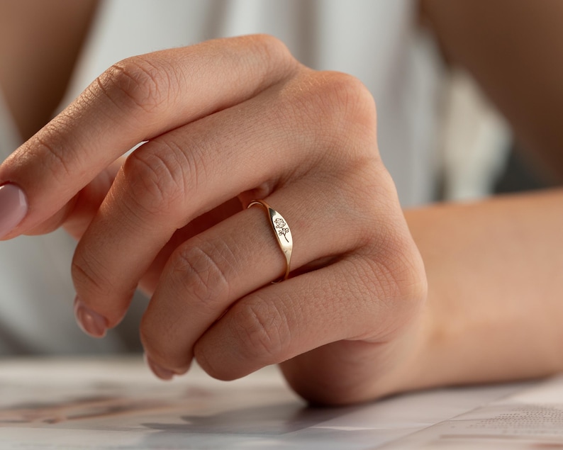 May include: A gold-toned signet ring featuring a small, engraved floral design on the flat, oval-shaped face. The band is thin and circular, with the metal surface displaying a polished finish that reflects light across the surface of the ring.
