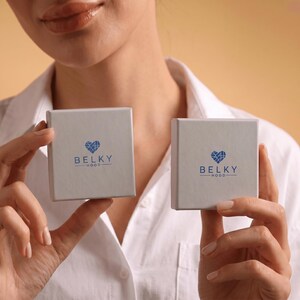 May include: Two square, light gray gift boxes featuring a blue logo with a heart-shaped icon composed of intersecting lines above the text BELKY HOOD. The boxes are held up, displaying the branding clearly on the smooth, matte-textured surfaces.