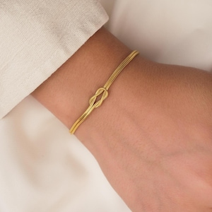 Può includere: Un braccialetto color oro con un design a nodo. Il braccialetto è realizzato in metallo sottile e strutturato e viene indossato su un polso. Lo sfondo è di un colore morbido e neutro.