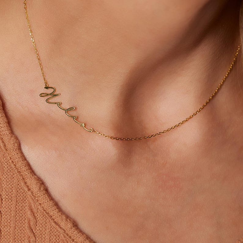 Pu&ograve; includere: Una collana d'oro con un ciondolo a forma di nome in corsivo. Il nome "Mela" &egrave; scritto in uno stile di scrittura delicato.