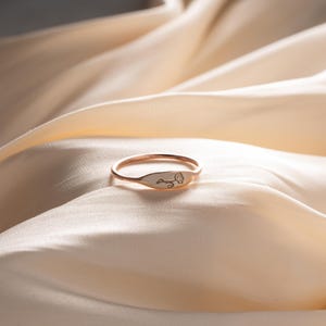 May include: A thin rose gold ring featuring a small, flat, oval-shaped face. The face of the ring is engraved with a delicate, minimalist line drawing of a single dandelion flower head with a curved stem, showing fine detail in the petals.