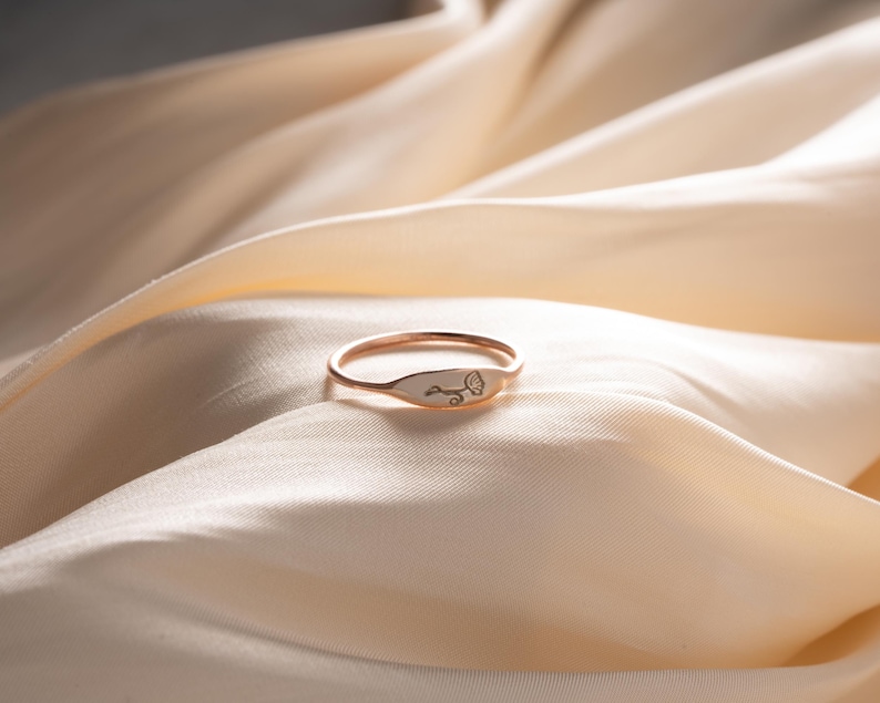 May include: A thin rose gold ring featuring a small, flat, oval-shaped face. The face of the ring is engraved with a delicate, minimalist line drawing of a single dandelion flower head with a curved stem, showing fine detail in the petals.
