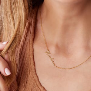Pu&ograve; includere: Una collana d'oro con un ciondolo a forma di nome in scrittura corsiva. Il ciondolo ha la forma della parola "Mamma".