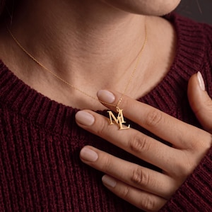 May include: A gold-toned necklace featuring a pendant with the capital letters M and L connected together. The pendant hangs from a delicate, thin gold-toned chain. The letters are rendered in a classic serif font with a smooth, polished finish.