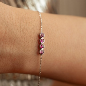 May include: A delicate silver bracelet featuring four round, vibrant red gemstones. The bracelet's chain is fine and elegant, with the gemstones set in individual bezels. The bracelet is designed to be worn on the wrist.