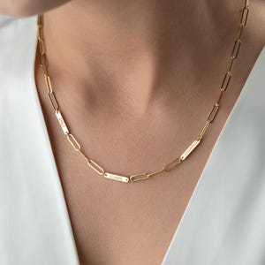 Peut inclure: Un collier chaîne doré avec des plaques nominatives rectangulaires. Les plaques nominatives sont gravées des noms "Emily", "Claire" et "Samantha". Le collier est porté autour du cou sur un fond blanc.