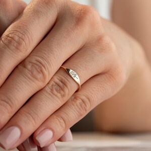 May include: A gold ring featuring a flat, elongated oval face engraved with a delicate, minimalist line drawing of a single rose. The band is thin and polished, showcasing a smooth, metallic finish that wraps around the finger.
