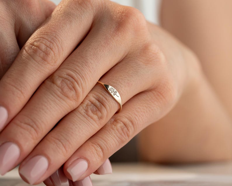May include: A gold ring featuring a flat, elongated oval face engraved with a delicate, minimalist line drawing of a single rose. The band is thin and polished, showcasing a smooth, metallic finish that wraps around the finger.