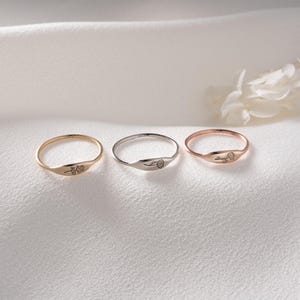 May include: Three thin metal rings arranged in a row, each featuring a small, delicate floral engraving on the flat front surface. The set includes one yellow gold-toned ring, one silver-toned ring, and one rose gold-toned ring, all with smooth bands.