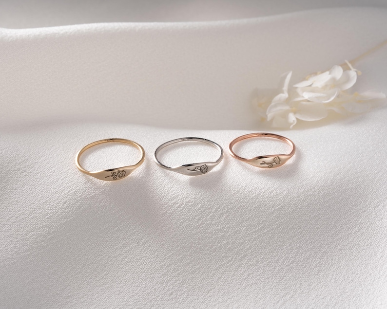 May include: Three thin metal rings arranged in a row, each featuring a small, delicate floral engraving on the flat front surface. The set includes one yellow gold-toned ring, one silver-toned ring, and one rose gold-toned ring, all with smooth bands.