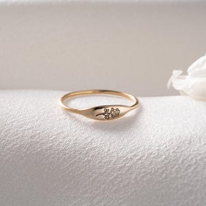 May include: A thin gold ring featuring a flat, oval-shaped face engraved with a delicate, single-line drawing of a rose. The band is slender and polished, showcasing a minimalist design with the floral engraving centered on the front surface.