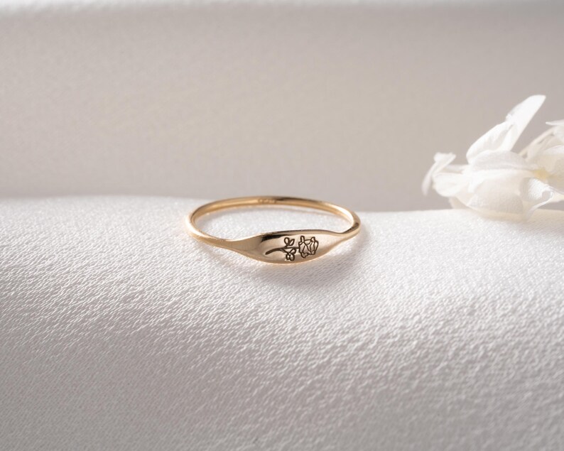 May include: A thin gold ring featuring a flat, oval-shaped face engraved with a delicate, single-line drawing of a rose. The band is slender and polished, showcasing a minimalist design with the floral engraving centered on the front surface.