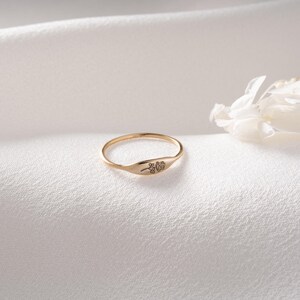 May include: A thin gold ring featuring a small, flat, oval-shaped face engraved with a delicate, minimalist line drawing of a single flower. The band is slender and polished, showcasing a warm metallic finish that catches the light.