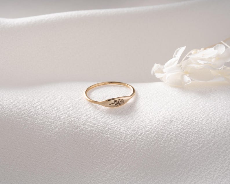 May include: A thin gold ring featuring a small, flat, oval-shaped face engraved with a delicate, minimalist line drawing of a single flower. The band is slender and polished, showcasing a warm metallic finish that catches the light.