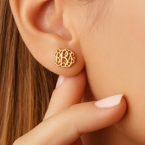 May include: A gold-colored monogram stud earring with an ornate, swirling design. The earring is affixed to an ear, showcasing the intricate details of the personalized initial design. The earring is a small, elegant piece of jewelry.