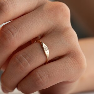 May include: A gold-toned ring featuring a flat, rectangular face engraved with a delicate, fine-line floral design. The band is thin and smooth, encircling the finger with a polished finish that reflects light across its metallic surface.