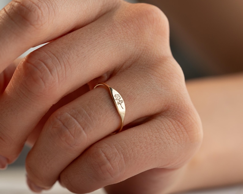 May include: A gold-toned ring featuring a flat, rectangular face engraved with a delicate, fine-line floral design. The band is thin and smooth, encircling the finger with a polished finish that reflects light across its metallic surface.