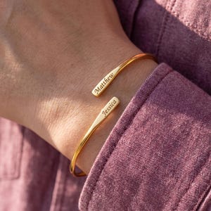 May include: A gold-toned open cuff bracelet featuring two flat, tapered ends. The top end is engraved with the name Matthew, and the bottom end is engraved with the name Jessica. The bracelet has a polished, smooth finish and a simple, curved design.
