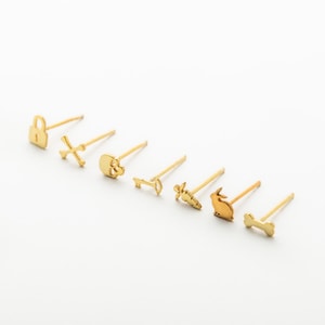 May include: A set of seven gold-toned stud earrings featuring distinct shapes including a padlock, an X, a skull, a key, a carrot, a rabbit, and a bone. Each earring has a thin post and is arranged in a row against a plain white background.