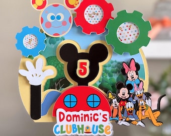 May include: A layered paper cake topper featuring a Mickey Mouse silhouette with the number 5, surrounded by colorful gears, a blue character with yellow ears, and a group of cartoon characters. The text reads Dominic's CLUBHOUSE.