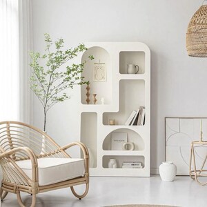 May include: A white modern bookshelf with arched openings and shelves, featuring a small potted plant, books, and decorative objects. A wicker chair with a white cushion sits in front of the bookshelf.