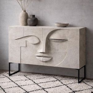 May include: A light-coloured, rectangular cabinet with a carved face design. The cabinet has black metal legs and is placed on a white rug with a black diamond pattern. Decorative vases and a bowl sit on top.