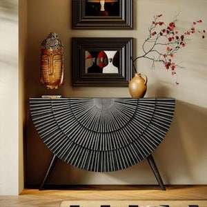 May include: A black and gray half-circle console table with a sunburst design. The table has three angled legs and is topped with a vase, a branch with red flowers, and a decorative mask. Two framed pictures hang on the wall above the table.