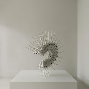 May include: A white, abstract sculpture with a spiral design, featuring numerous pointed protrusions. The sculpture is placed on a white, rectangular pedestal in a minimalist setting. The piece has black dot details.