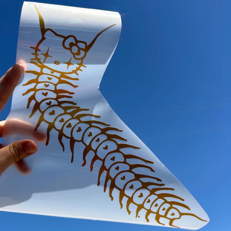 May include: A gold vinyl decal of a Hello Kitty-inspired centipede with a pink bow. The centipede has a long body with many legs and small hearts along its body.