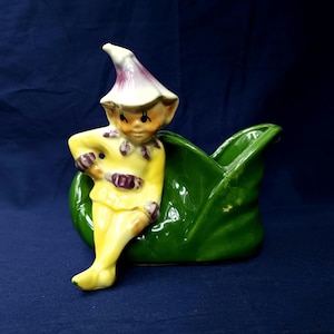 May include: A vintage ceramic planter featuring a whimsical elf figure in a yellow outfit with purple accents, leaning against a large, glossy green leaf-shaped container. The elf has a white hat with a purple tip. The planter is likely for small plants or flowers.